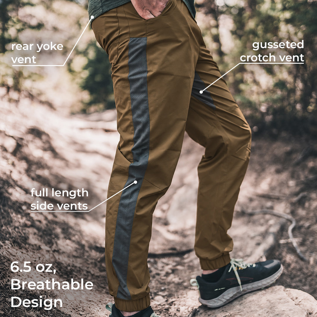 A person outdoors wearing brown hiking pants with dark gray side vents and text callouts pointing to rear yoke vent, gusseted crotch vent, full length side vents, and 6.5 oz breathable design.