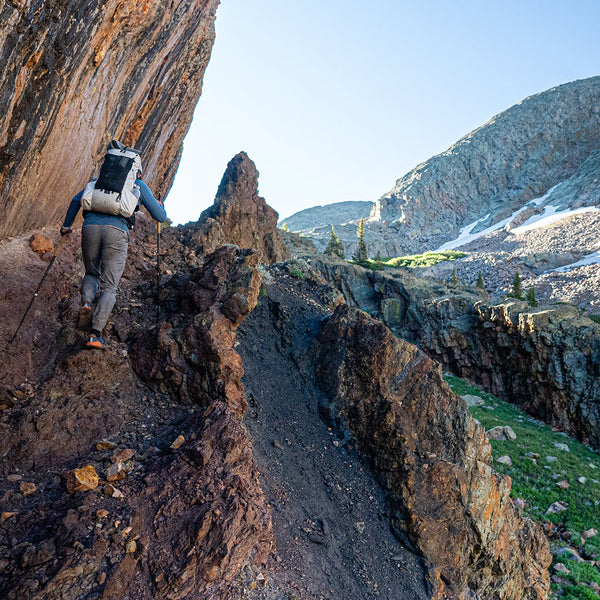Back view of a hiker using trekking poles to navigate a steep, rocky cliffside path while carrying the Outdoor Vitals CS40 Ultra Backpack in high-elevation terrain.
