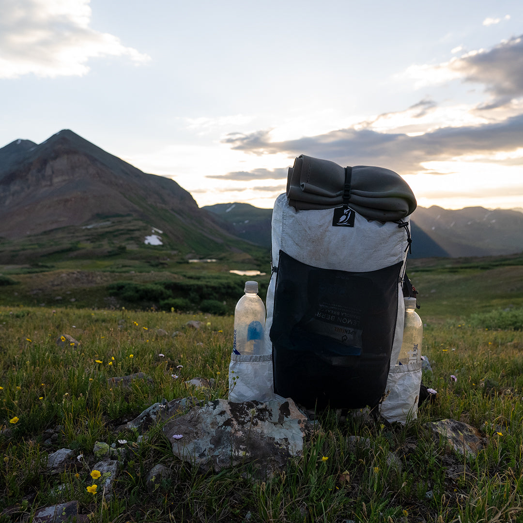 Outdoor Vitals CS40 Ultra Backpack fully packed with water bottles in the side pockets and gear strapped to the top, resting on a grassy alpine ridge with mountains in the background.