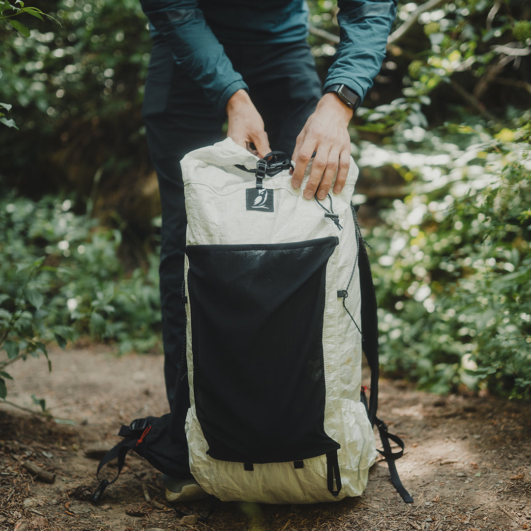 Front view of a hiker adjusting the top clip closure of the Outdoor Vitals CS40 Ultra Backpack on a wooded dirt trail, highlighting the front stretch mesh pocket and side compression straps.