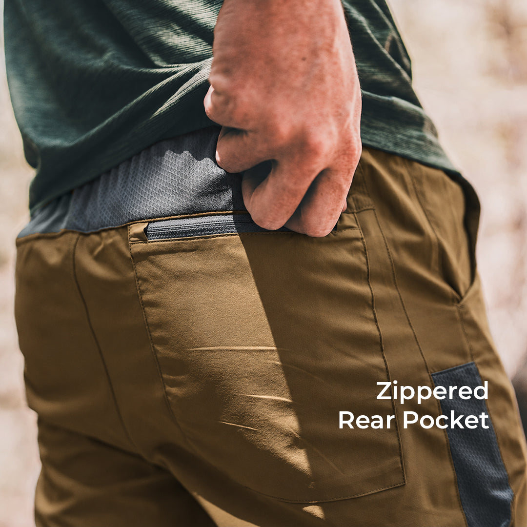 Close-up of a person wearing brown pants, showing the back pocket with a zipper and the text 'Zippered Rear Pocket' on a blurred outdoor background.