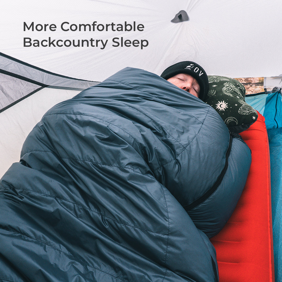 A person sleeping comfortably inside a tent, wrapped in a dark blue sleeping bag with a patterned pillow. Text in the upper left reads 'More Comfortable Backcountry Sleep'.