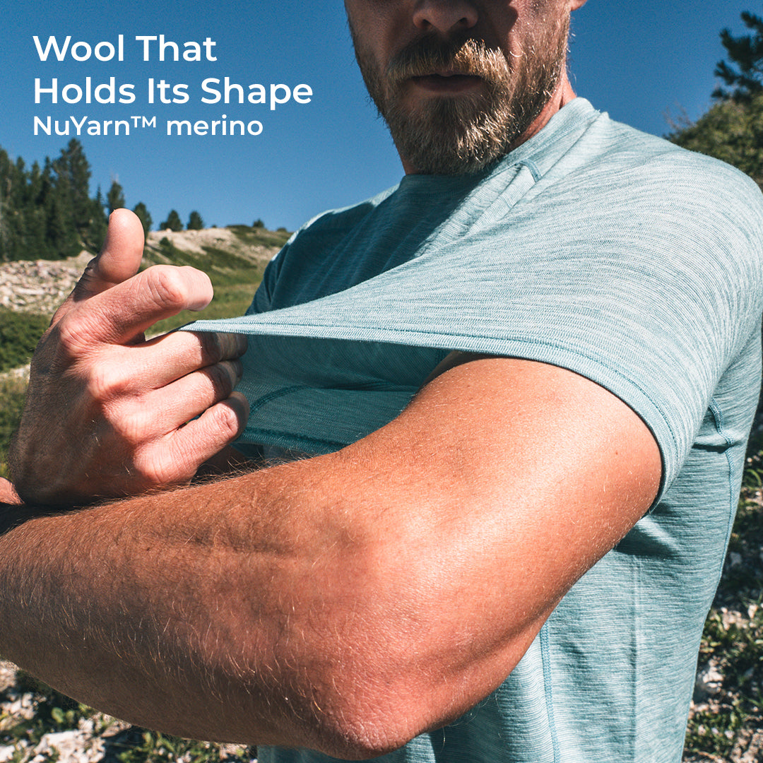 A man outdoors wearing a light merino wool t-shirt and stretching it to show its elasticity. Text in the top left reads 'Wool That Holds Its Shape NuYarn™ merino'.