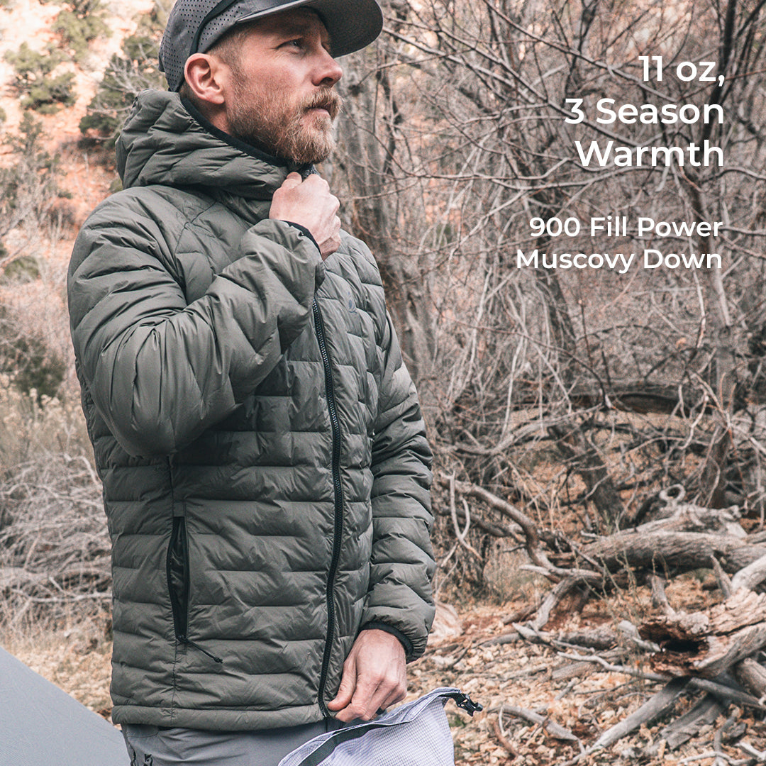 A man wearing a gray puffer jacket and a black cap stands outdoors in a wooded setting. Text on the right reads '11 oz, 3 Season Warmth' and '900 Fill Power Muscovy Down'.