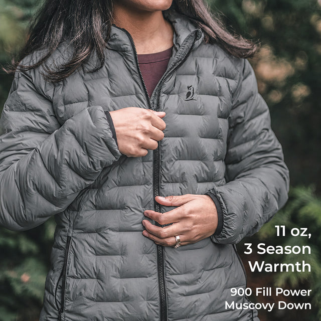 A woman wearing a dark gray quilted puffer jacket, shown from the chest down, with text overlays on the right detailing its features: '11 oz, 3 Season Warmth, 900 Fill Power Muscovy Down'.