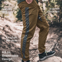 A person outdoors wearing brown hiking pants with dark gray side vents and text callouts pointing to rear yoke vent, gusseted crotch vent, full length side vents, and 6.5 oz breathable design.