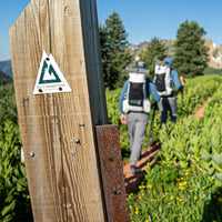 Wooden trail marker post with a Colorado Trail sign in the foreground, with two hikers wearing Outdoor Vitals CS40 Ultra Backpacks walking down a scenic mountain trail in the background.