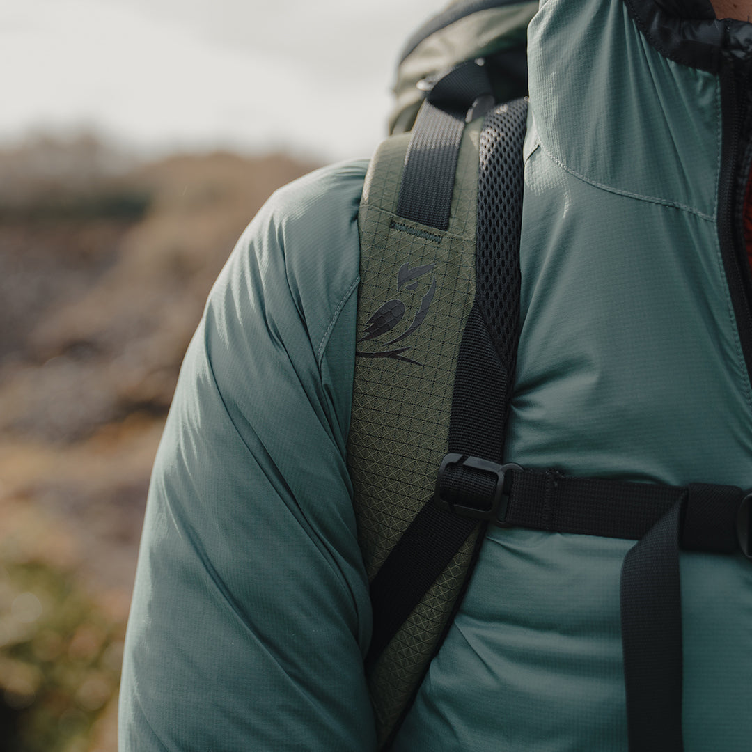 Close-up view of the padded shoulder strap on the Outdoor Vitals KotaUL Ultralight Travel & Adventure Backpack, showcasing the printed bird logo, ripstop fabric, breathable mesh padding, and adjustable sternum strap buckle.