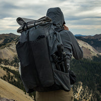 Hiker wearing a Outdoor Vitals Shadowlight backpack with gear strapped to the top and tripod stored in the side pocket, overlooking a rugged mountain ridgeline under cloudy skies.