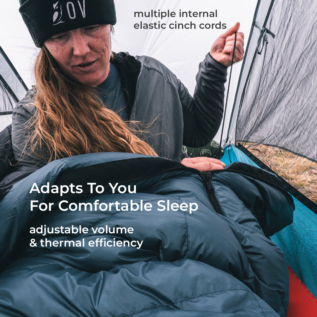A person inside a tent wearing a black beanie with white text and a dark gray hoodie, adjusting a dark blue sleeping bag. White text overlays describe the product features: 'multiple internal elastic cinch cords' and 'Adapts To You For Comfortable Sleep adjustable volume & thermal efficiency'.