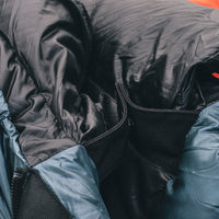Close-up detail of a backpacking sleeping bag showing the black insulated fill, black zipper track, black snap closure at the collar, and blue-gray shell fabric.