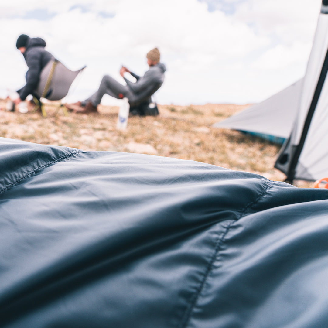 A close-up of a dark blue sleeping bag  in the foreground, with a blurred outdoor camping scene in the background showing two people sitting near a tent on a rocky hillside.