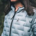 A close-up of a person wearing a light blue puffer jacket outdoors. The jacket has a black zipper and a small black bird logo on the left chest.