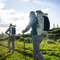 Two hikers using trekking poles and wearing Outdoor Vitals CS40 Ultra Backpacks and Satu Adventure Pants while trekking along a sunlit mountain trail surrounded by wildflowers.