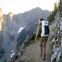 Back view of a hiker carrying the Outdoor Vitals CS40 Ultra Backpack while trekking along a narrow mountain cliff path overlooking a deep valley.