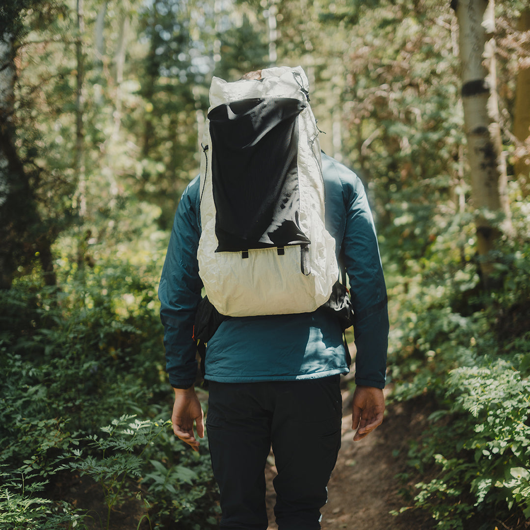 Back view of a hiker walking along a wooded trail carrying the Outdoor Vitals CS40 Ultra Backpack, highlighting the large front stretch pocket and ergonomic fit.