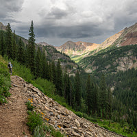 Distant view of a backpacker carrying the Outdoor Vitals CS40 Ultra Backpack while hiking along a rocky mountain ridge trail with pine trees and peak landscape views.