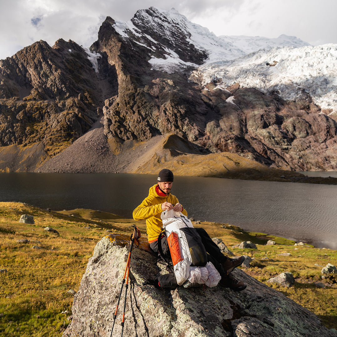 A hiker seated on a large boulder adjusting the top closure of the Outdoor Vitals CS40 Ultra Backpack, set against an alpine lake and massive snow-covered mountain backdrop.