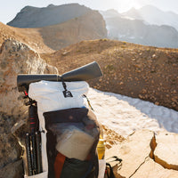 Close-up view of the Outdoor Vitals CS40 Ultra Backpack resting against a rock in a high-alpine landscape, fully loaded with a sleeping pad strapped on top, tripod attached to the side, and gear packed in the front mesh pocket.