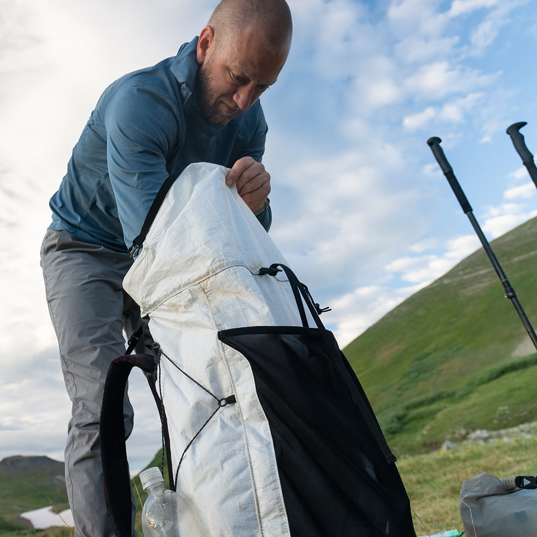 Hiker reaching into the open main compartment of the Outdoor Vitals CS40 Ultra Backpack outdoors, with trekking poles and mountain slopes visible in the background.