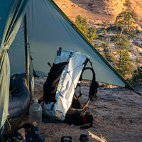 The Outdoor Vitals CS40 Ultra Backpack standing upright under the vestibule of a tarp shelter at a desert campsite, surrounded by hiking gear and overlook views.