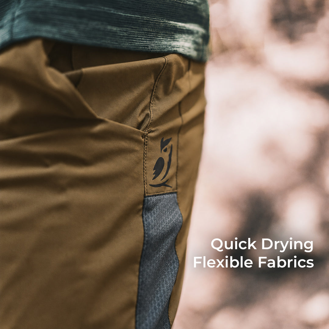 Close-up of a person wearing brown and gray outdoor pants with a dark logo patch on the pocket, set against a blurred outdoor background with text reading 'Quick Drying Flexible Fabrics'.