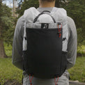Direct rear view of a hiker wearing the Outdoor Vitals Skyline 30 Fastpack, displaying the large front stretch-mesh pocket, side bottle pockets, brand logo, and roll-top compression strap system.