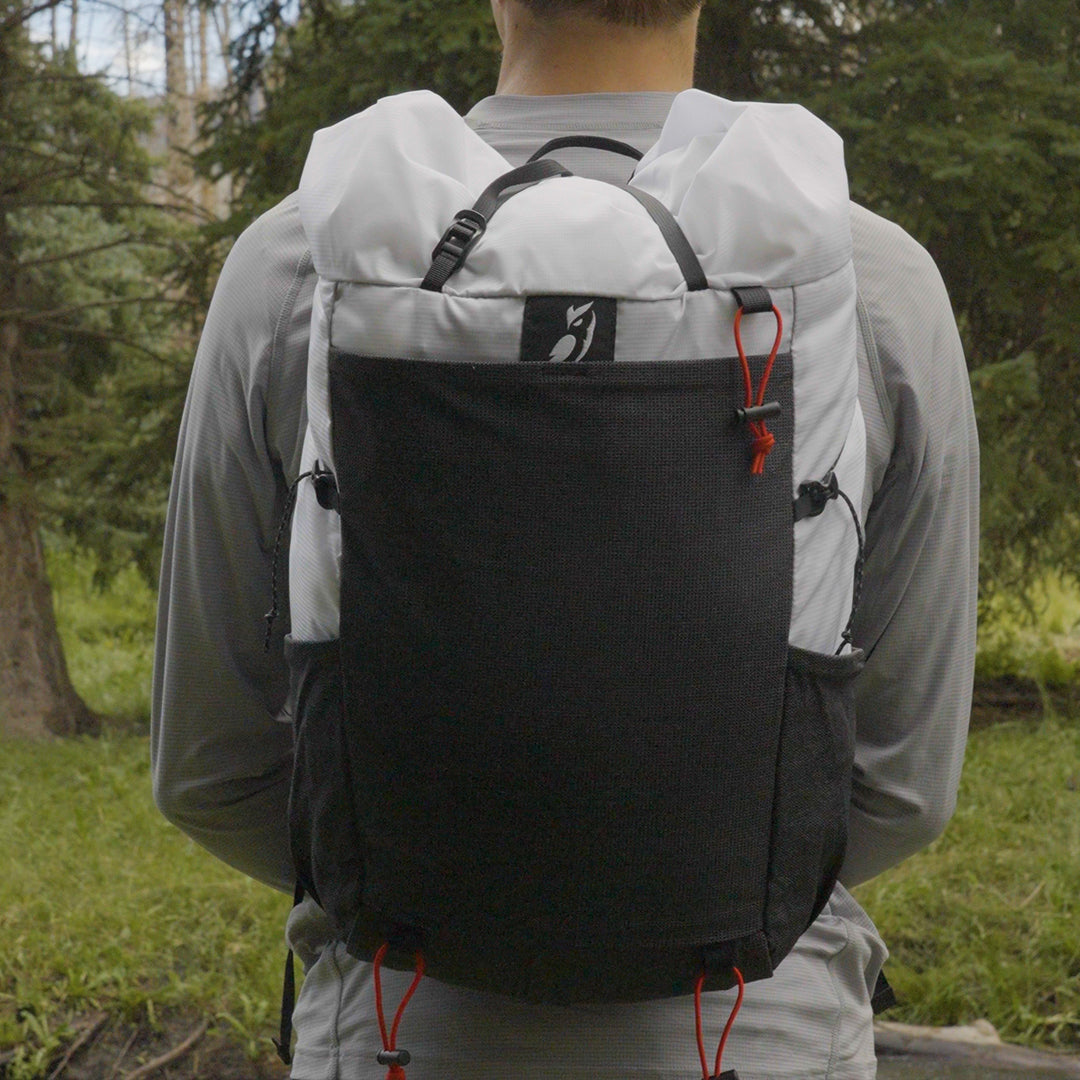 Direct rear view of a hiker wearing the Outdoor Vitals Skyline 30 Fastpack, displaying the large front stretch-mesh pocket, side bottle pockets, brand logo, and roll-top compression strap system.