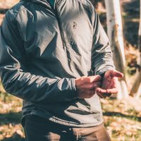 A man wearing a gray lightweight zip-up jacket outdoors in a wooded setting, while he adjusts his elastic cuffs.