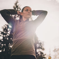 A woman outdoors with her hands behind her head adjusting her ponytail, wearing a light brown quarter-zip athletic pullover and dark pants, set against a bright, sunlit sky and blurred trees.