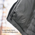 Close-up of a dark gray quilted puffer jacket corner with a black zipper and black plastic adjuster, set against a blurred outdoor background with text reading 'Drop Back Hem + Internal Adjusters'.