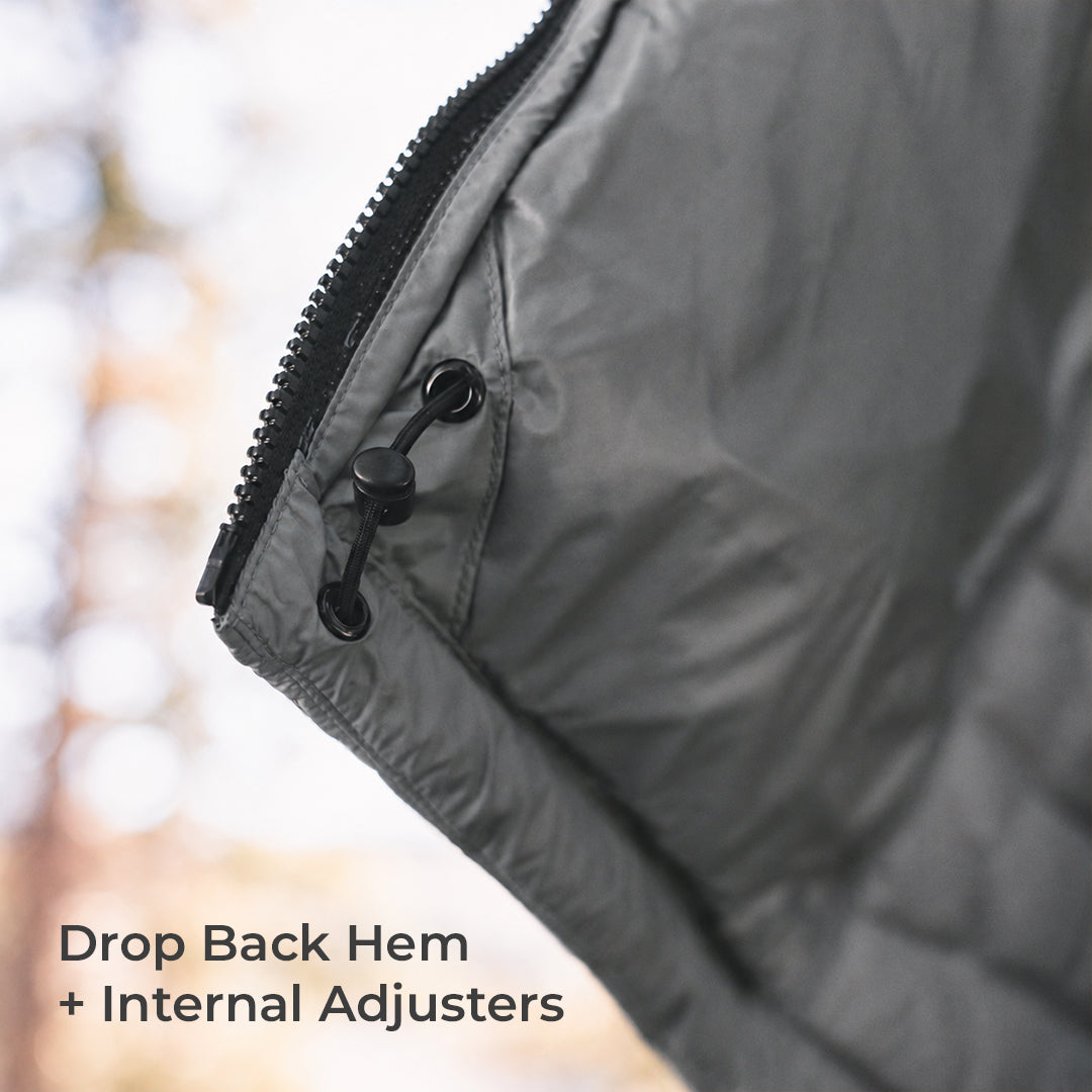Close-up of a dark gray quilted puffer jacket corner with a black zipper and black plastic adjuster, set against a blurred outdoor background with text reading 'Drop Back Hem + Internal Adjusters'.