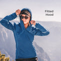 A woman wearing a fitted blue hooded athletic jacket stands outdoors against a blurred mountain background, adjusting the hood. The text 'Fitted Hood' appears in the upper right.