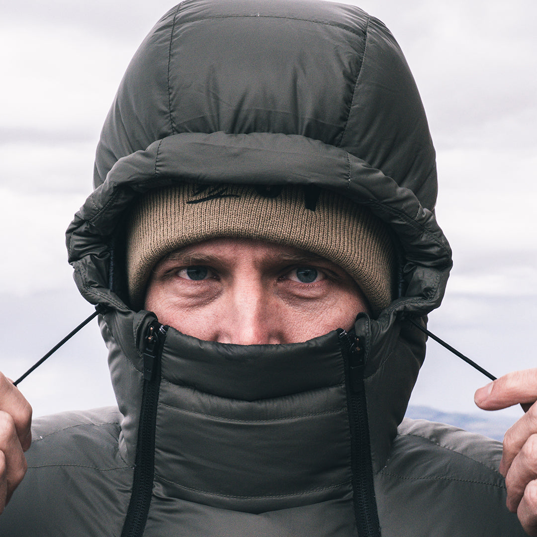 A close-up of a man wearing a dark  puffer jacket with an integrated hood and face warmer, paired with a tan beanie, against an overcast sky.