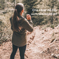 A woman standing on a dirt trail in a forest, viewed from behind, wearing an olive green hooded jacket and black leggings. White text on the right reads 'One Layer All Day' and 'comfortable from 25-50F'.