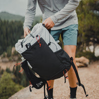 A hiker holding the Outdoor Vitals Skyline 30 Fastpack by its top loop, highlighting the front stretch mesh pocket, shoulder harness water bottle sleeve, and roll-top closure.