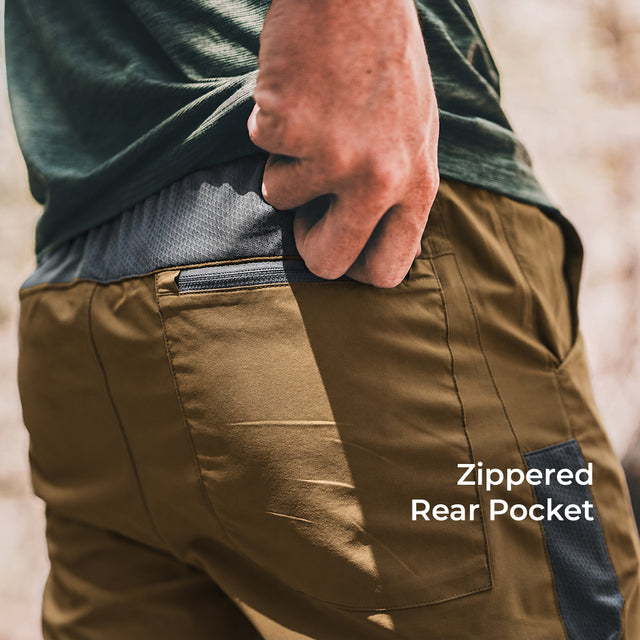 Close-up of a person wearing brown pants, showing the back pocket with a zipper and the text 'Zippered Rear Pocket' on a blurred outdoor background.