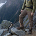 Close-up of a hiker standing on rocky terrain wearing Outdoor Vitals Satu Adventure Pants, hiking boots, and a puffer jacket with a mountain backdrop.