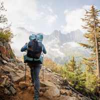 Rear view of a hiker ascending a rugged mountain trail carrying the Outdoor Vitals Shadowlight Carbon 60 Backpack, featuring large rear mesh storage and side water bottle pockets.