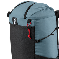 Close-up of a backpack with a dusty blue top section, a black lower pocket, black straps, and a red drawcord detail on a white background.