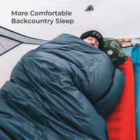 A person sleeping comfortably inside a tent, wrapped in a dark blue sleeping bag with a patterned pillow. Text in the upper left reads 'More Comfortable Backcountry Sleep'.
