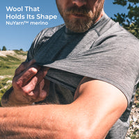 A close-up outdoor shot of a bearded man lifting his short-sleeve shirt to show the fabric shape retention and stretch. Text in the top left reads 'Wool That Holds Its Shape NuYarn™ merino'.
