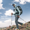 A man hiking on a rocky mountain under a blue sky with clouds, wearing a hooded long-sleeve shirt, hiking pants, and carrying a black backpack with trekking poles. White text on the left reads 'Naturally Odor Resistant'.