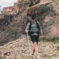 A man hiking on a rocky mountain slope using trekking poles, wearing a long-sleeve top, shorts, and a backpack.