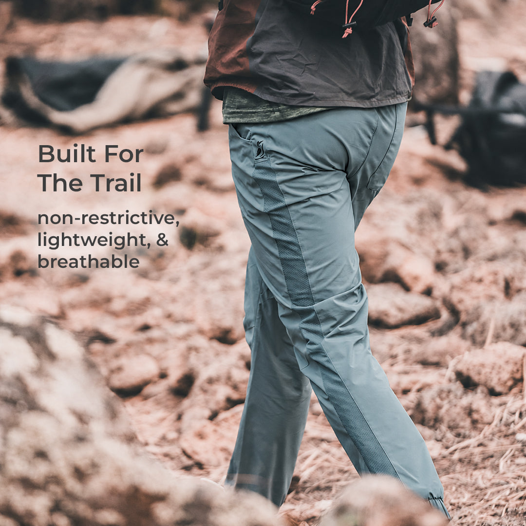 A person outdoors on a rocky trail wearing light blue-gray hiking pants and a dark jacket. Text on the left reads, 'Built For The Trail non-restrictive, lightweight, & breathable.