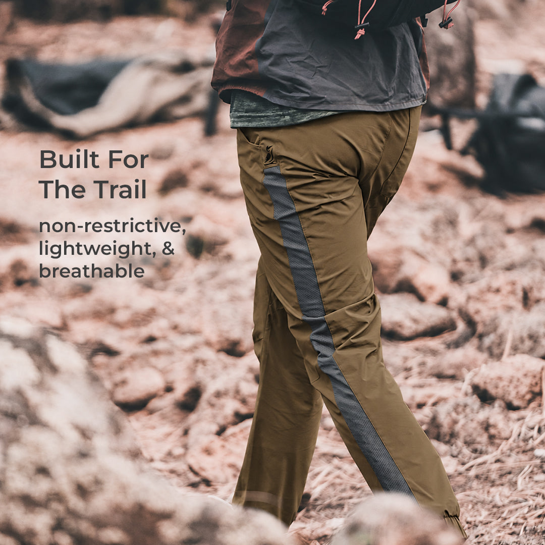 A person outdoors wearing brown hiking pants with a dark gray side panel, standing on rocky terrain. Text on the left reads 'Built For The Trail' and 'non-restrictive, lightweight, & breathable'.