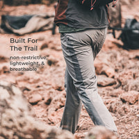 A person wearing light gray hiking pants with dark side panels walks through rocky, reddish-brown outdoor terrain. Text on the left reads, 'Built For The Trail' and 'non-restrictive, lightweight, & breathable.