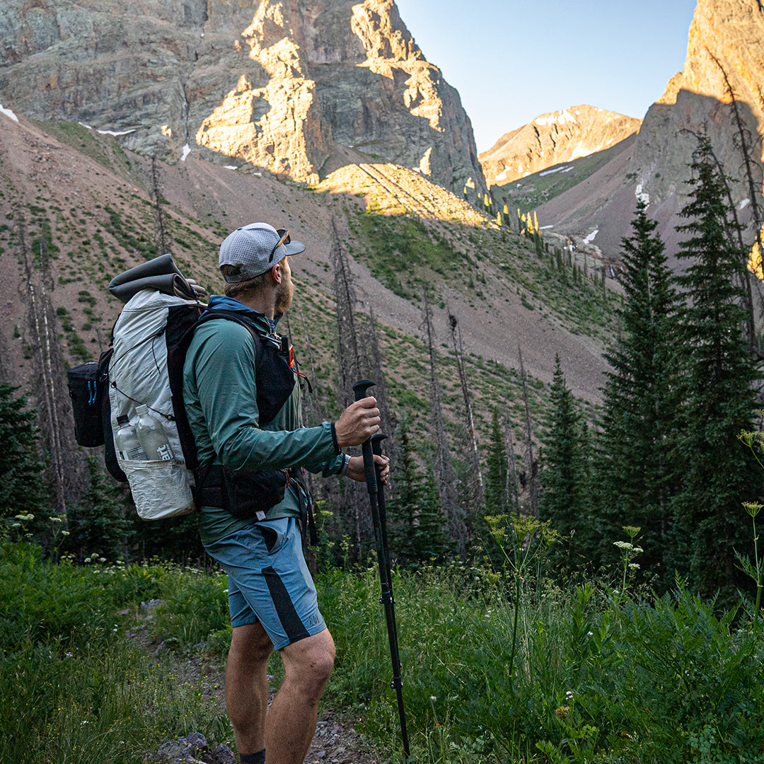 Side view of a male hiker holding trekking poles and looking up at steep mountain peaks while wearing the Outdoor Vitals CS40 Ultra Backpack with side water bottle pockets and attached gear.