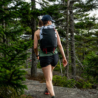 Back view of a female hiker walking through a dense pine forest wearing the Outdoor Vitals Skyline 30 Fastpack, showcasing the large stretch-mesh pocket and roll-top design in an outdoor setting.