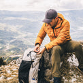 A man in an orange puffer jacket and dark cap sits on a rocky mountain ridge, organizing gear in a light gray and black hiking backpack. The background shows a vast, hazy mountain valley under a cloudy sky.